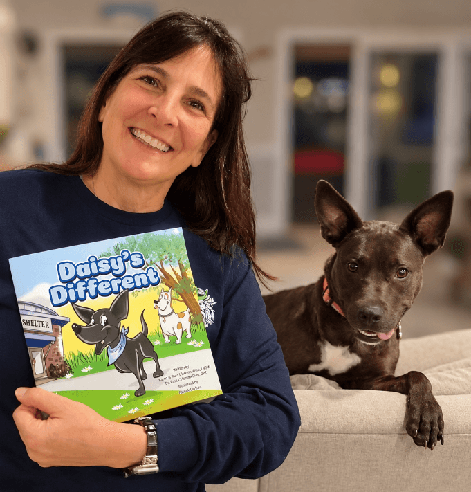 Karen smiling and holding a copy of Daisy's Different, with Daisy resting beside her.